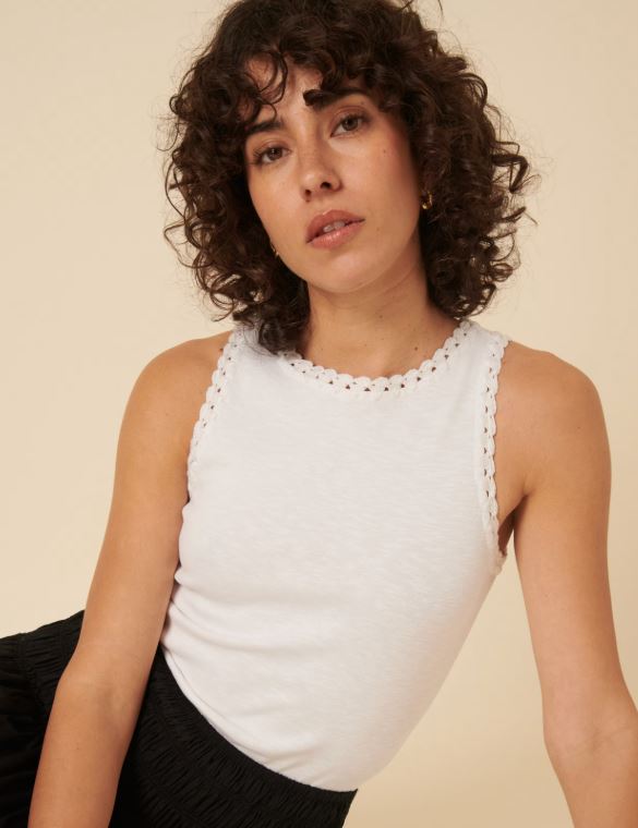 Woman with curly brown hair wearing a sleeveless white top with scalloped edges and a black skirt, posing against a beige background.