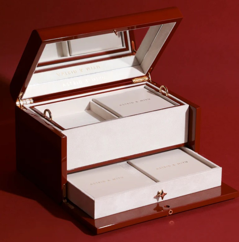 Astrid & Miyu 14K Solid Gold Advent Calendar 2025 A glossy brown jewelry box with a mirror in the lid, containing several rectangular cream-colored boxes labeled "ASTRID & MIYU" on a red background.