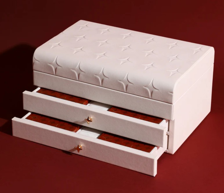 Astrid & Miyu 24-Day Advent Calendar 2025 A rectangular, upholstered white jewelry box with a quilted pattern on the lid and two open drawers lined in red, set against a burgundy background.