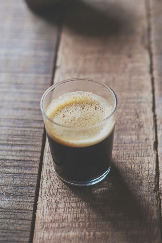 A clear glass filled with dark espresso and a layer of crema sits on a rustic wooden surface.