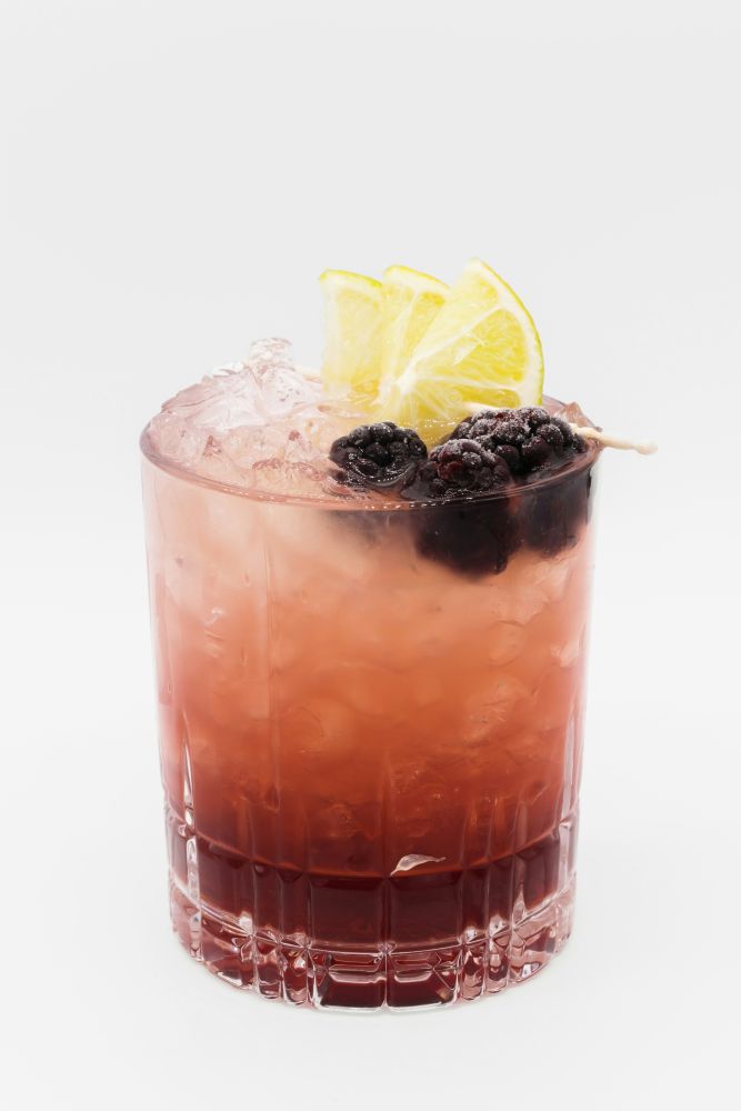 A glass of pink cocktail with ice, garnished with two lime wedges and blackberries, against a plain white background.