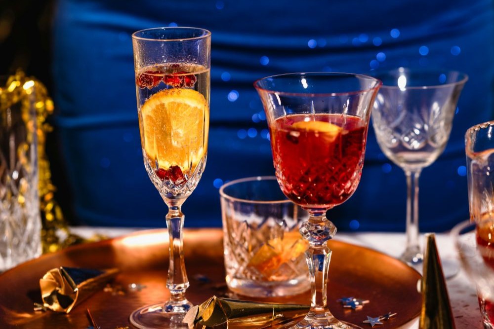 Two glasses with festive drinks—one with orange and cranberries, the other with lemon—sit on a copper tray amid party decor, perfect for easy Christmas cocktails or anyone seeking easy cocktails to make at home.