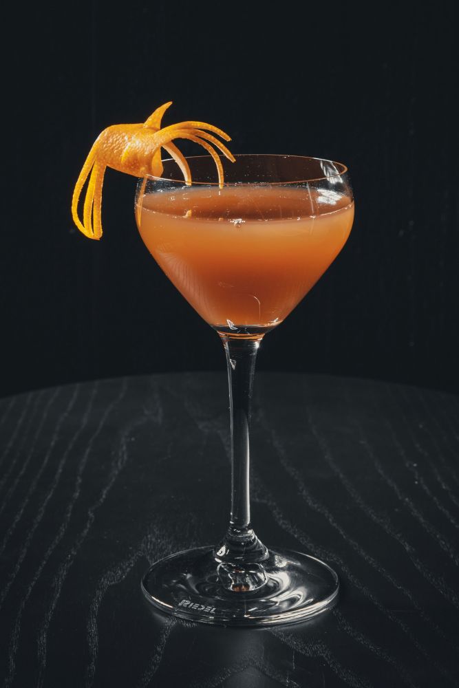 A cocktail in a stemmed glass garnished with an elaborate orange peel twist, set against a dark background.