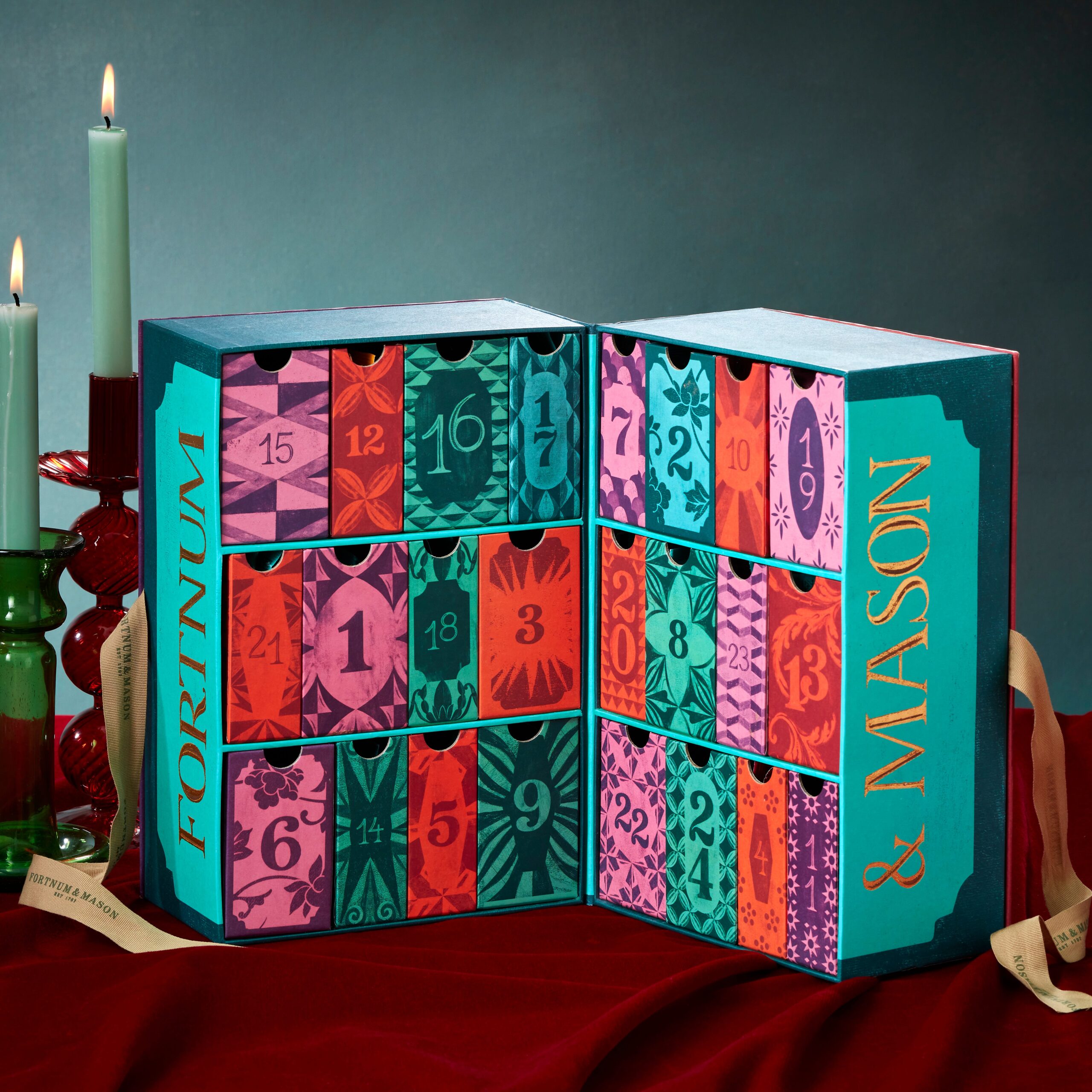 An open Fortnum & Mason advent calendar with numbered, colorful drawers sits on a red cloth next to green and red candles.