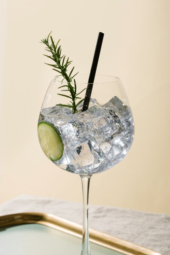 A cocktail glass filled with clear liquid, ice cubes, a slice of lime, a sprig of rosemary, and a black straw, placed on a tray.