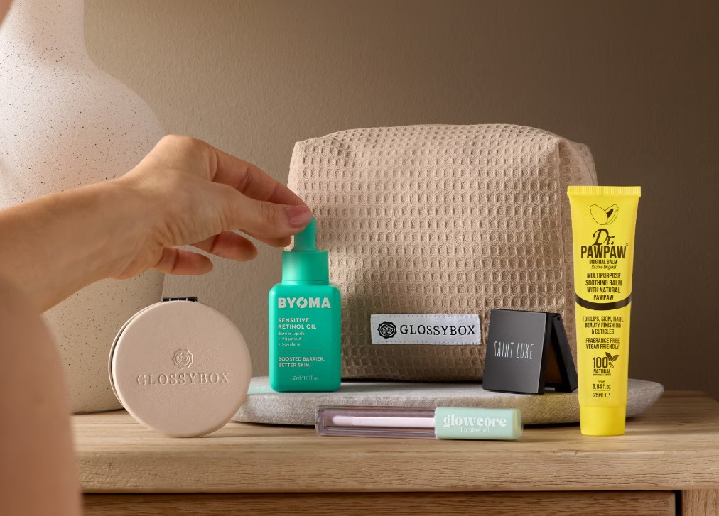 A hand holds a bottle of BYOMA serum among various skincare and cosmetic products, including a washbag labeled “Glossybox,” on a wooden surface.