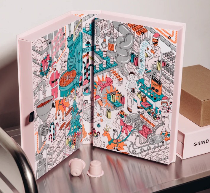 An illustrated advent calendar with colorful drawings of coffee-related scenes is propped open on a counter next to coffee pods and boxes.