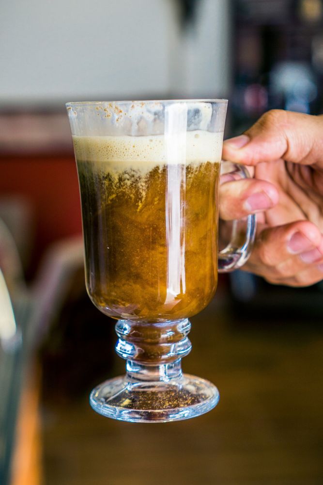 A hand holds a clear glass mug filled with a dark coffee drink topped with a creamy foam layer.