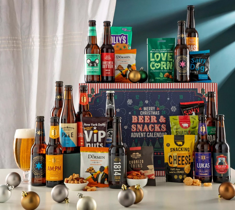 A selection of beers and snacks are arranged around a Christmas-themed advent calendar box, with a poured beer glass and festive ornaments in the foreground.