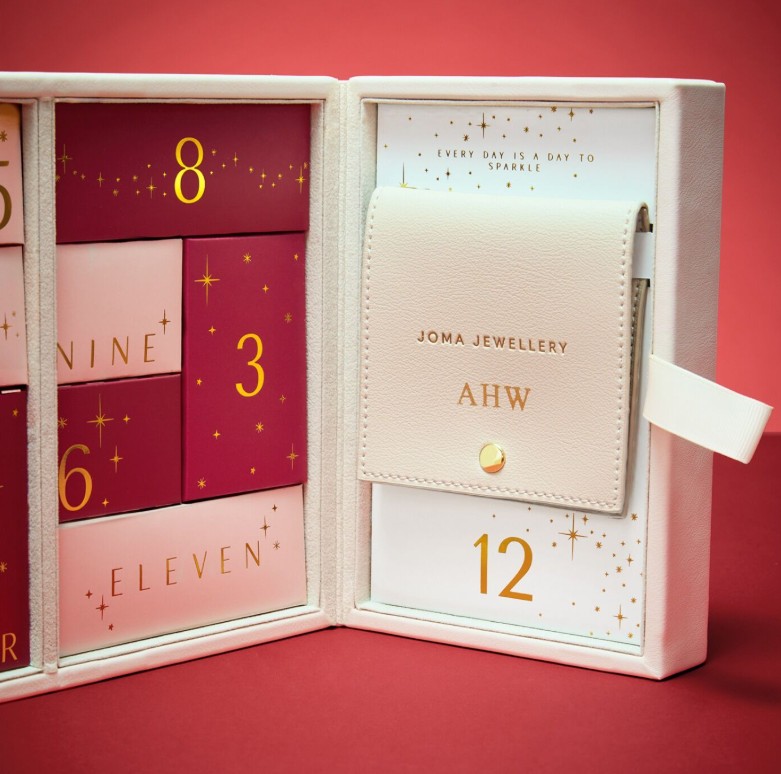 Joma 12 Days of Sparkle Advent Calendar in Gold Plating A white and gold jewelry advent calendar box is open, revealing numbered compartments and a cream-colored pouch labeled "Joma Jewellery" and "AHW" on a red background.