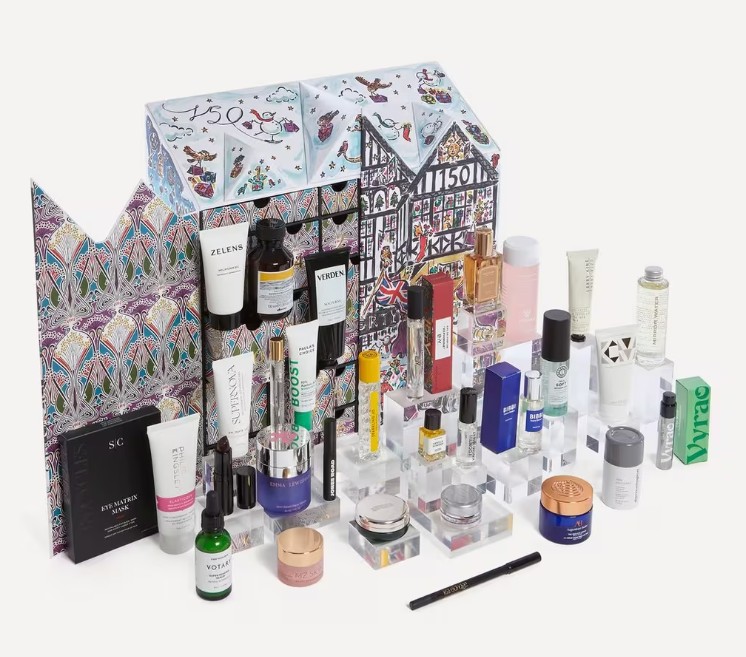 A variety of skincare and beauty products displayed in front of an open, decorative advent calendar featuring colorful, house-shaped packaging.