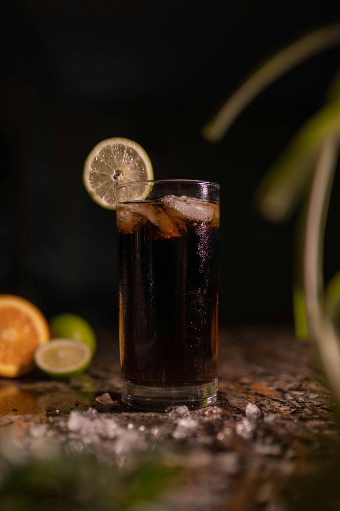 A glass of dark soda with ice and a lime slice garnish sits on a marble surface, with citrus fruits and ice scattered in the background.