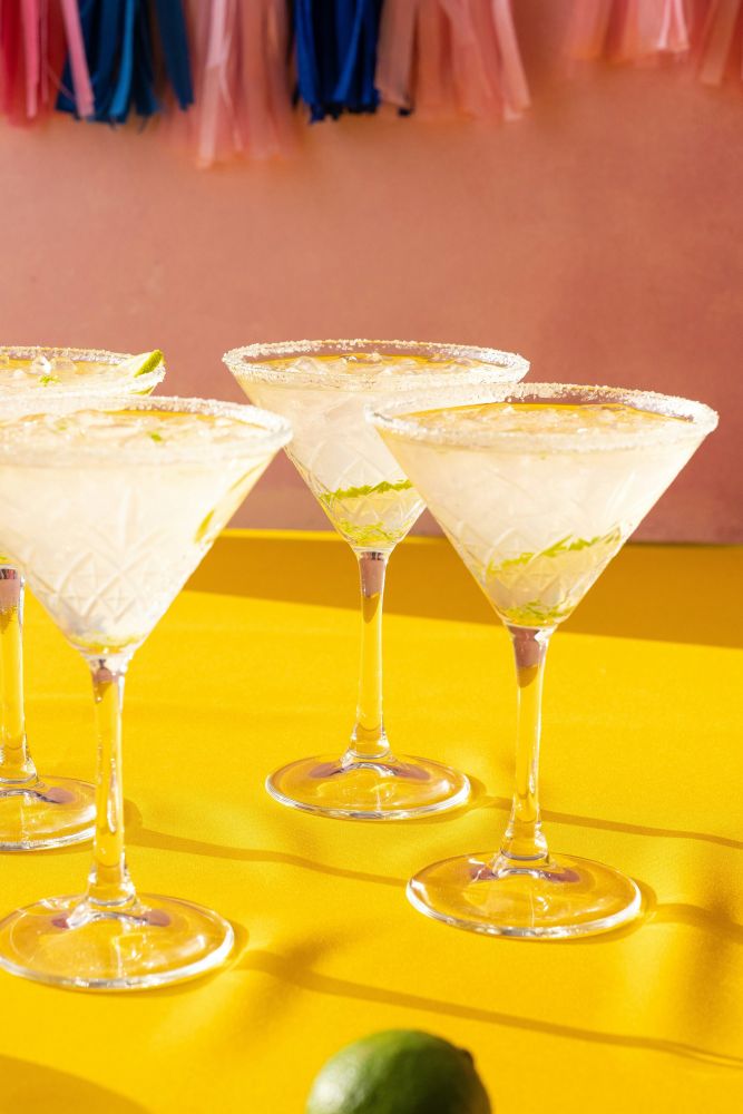 Four margarita glasses with salt rims and lime slices are placed on a yellow surface, with a pink wall and colorful tassels in the background.