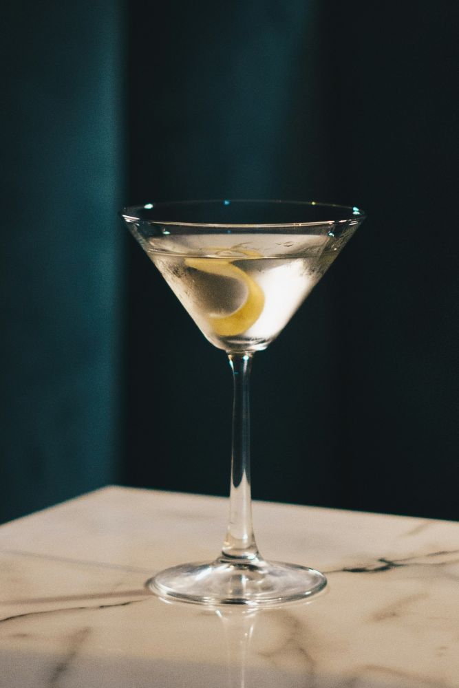 A clear martini in a stemmed glass with a lemon twist garnish, placed on a marble surface against a dark background.