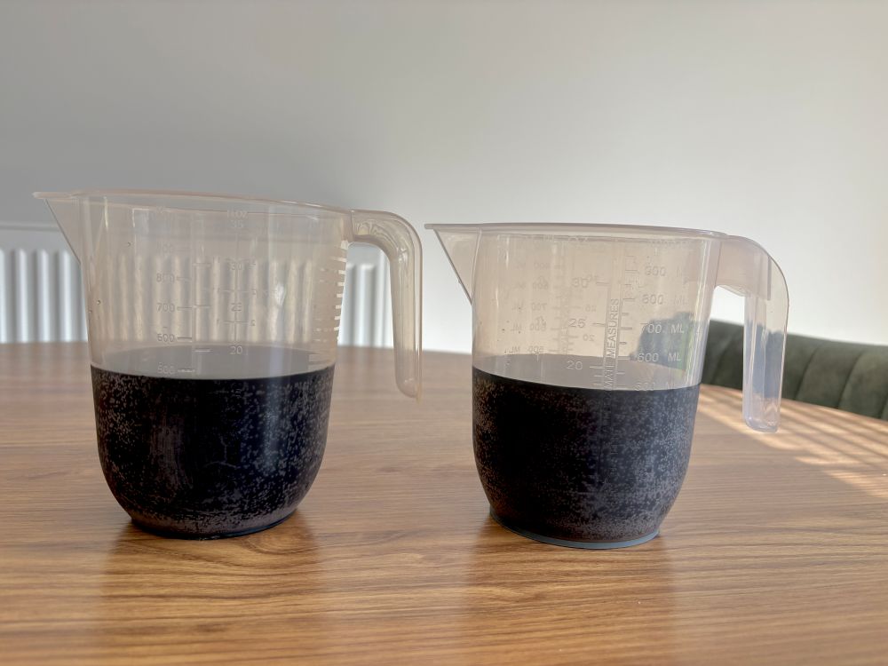 Two clear plastic measuring jugs filled with a dark liquid sit side by side on a wooden table. Each jug is marked with measurement lines.