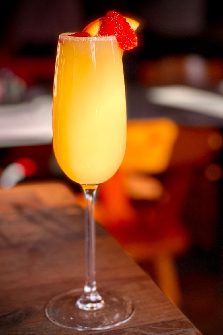 A champagne flute filled with a yellow-orange drink, garnished with a sliced strawberry on the rim, placed on a wooden table.