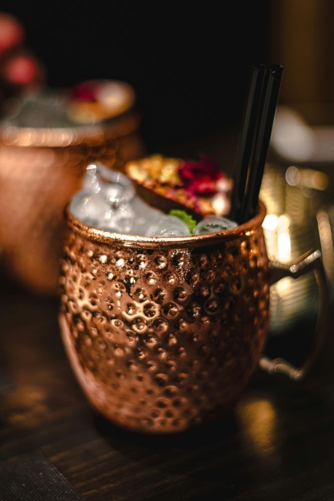A copper mug filled with ice, garnished with fruit and flowers, and two black straws, sitting on a dark table.