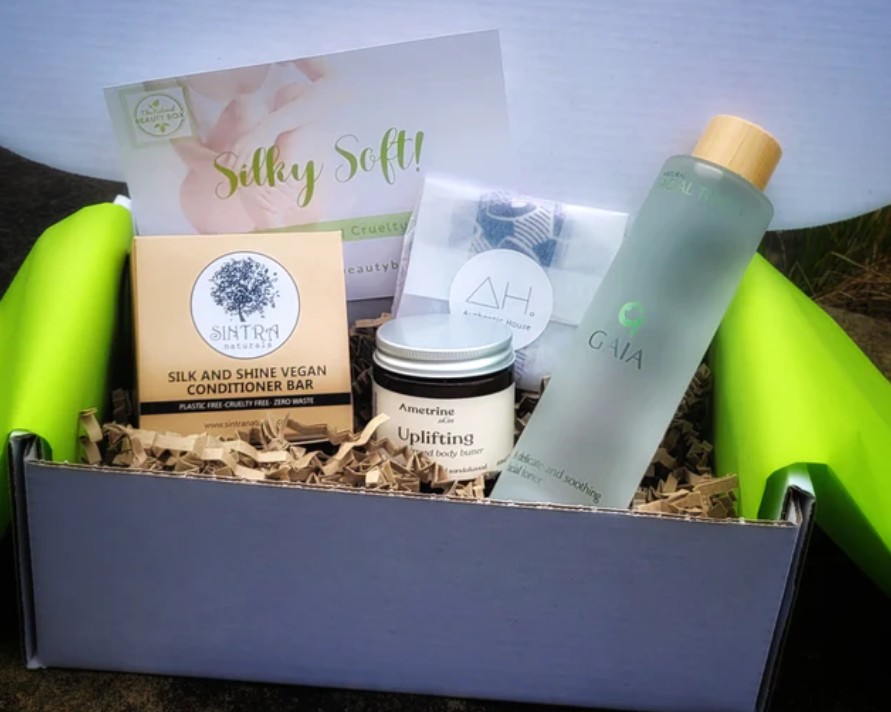 A gift box containing a conditioner bar, body butter, facial rounds, a bottle of liquid, and an informational card, arranged with shredded paper filler.