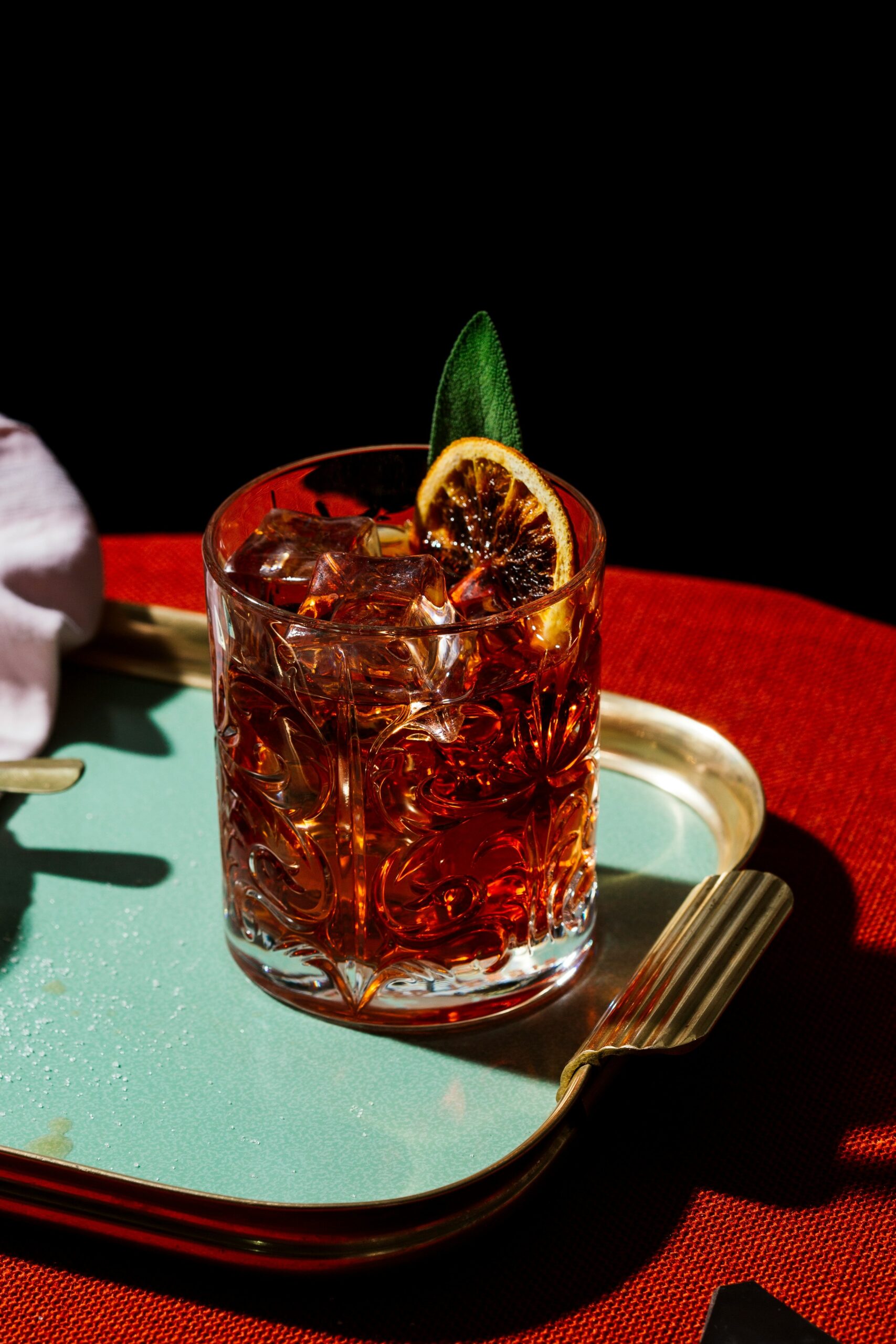 A glass of iced cocktail garnished with a dried orange slice and green leaf sits on a gold-edged tray against a dark background.