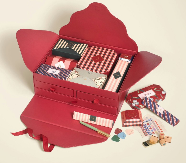 A red gift box is open, displaying various stationery items, envelopes, pens, stickers, cards, and small boxes with decorative patterns arranged neatly inside.
