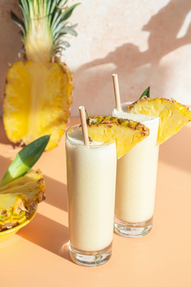 Two tall glasses of pineapple smoothie with large pineapple wedges and straws, set on a peach-colored surface with a halved pineapple in the background.