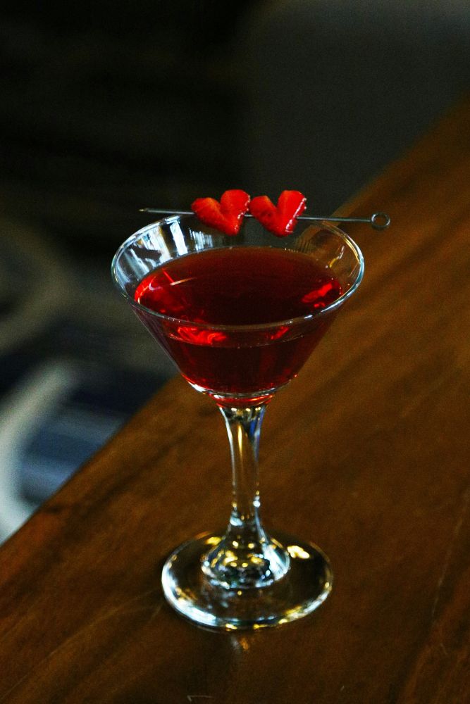 A red cocktail in a martini glass on a wooden surface, garnished with two heart-shaped fruit pieces on a skewer.