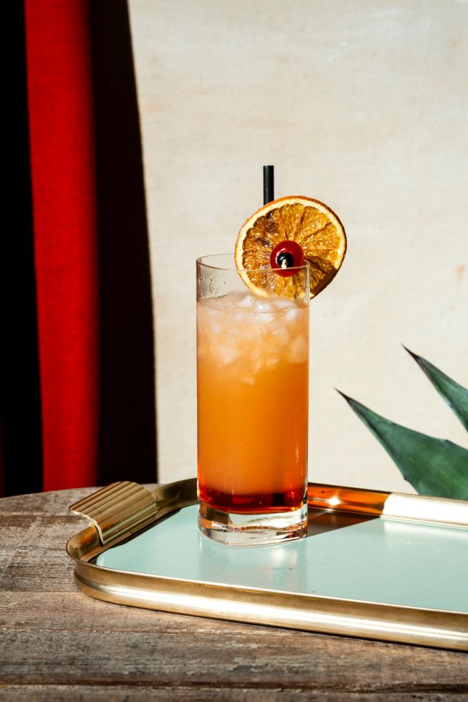 A tall glass of orange cocktail with ice, garnished with a dried orange slice and cherry, on a tray with a straw, placed on a wooden table.