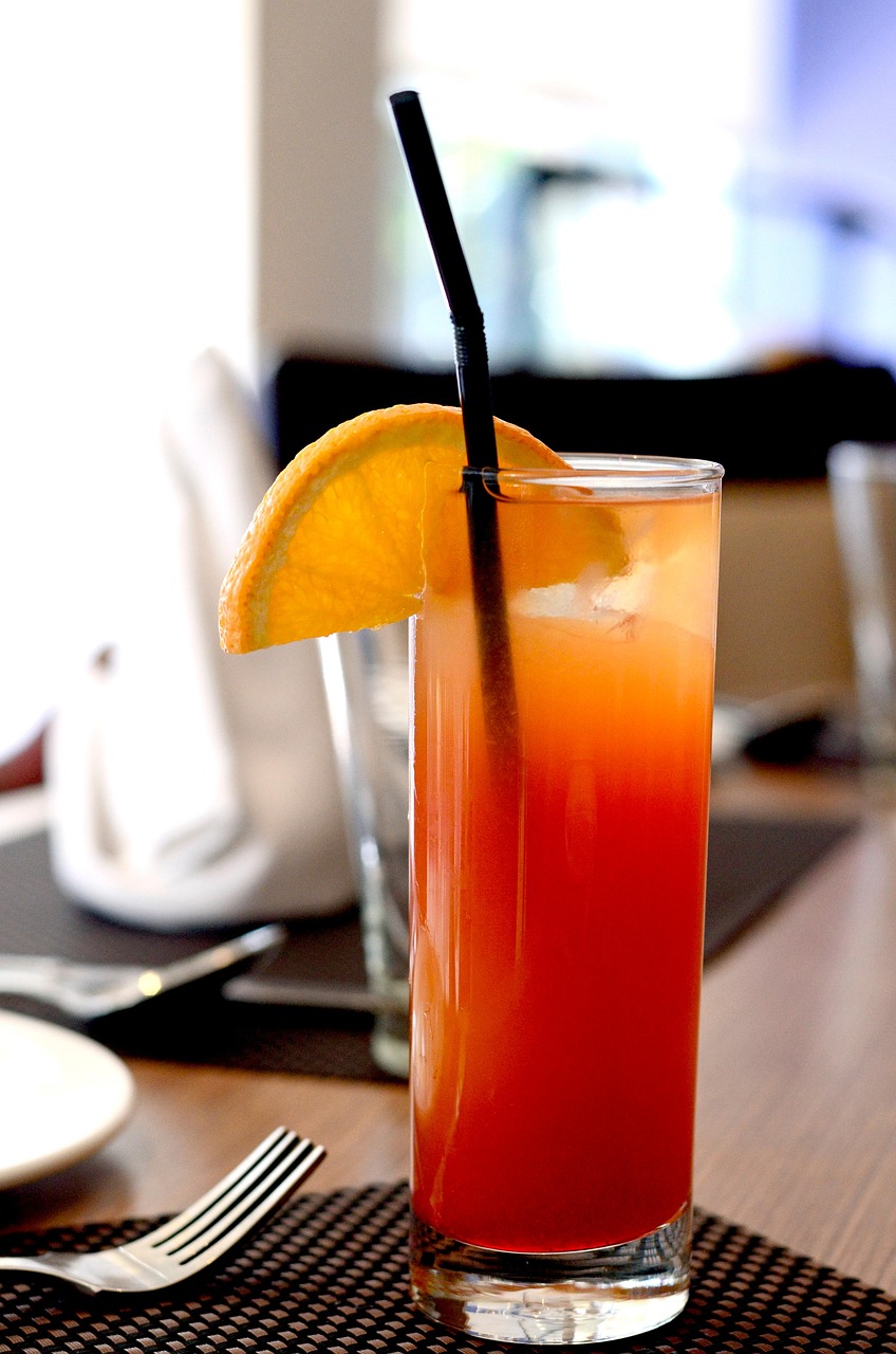 A tall glass of orange and red cocktail with ice, garnished with an orange slice and a black straw, placed on a table setting.