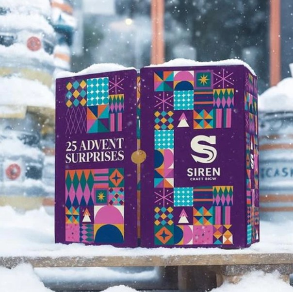 A colorful Siren Craft Brew advent calendar box labeled "25 Advent Surprises" sits on snow, with kegs and a window in the background.