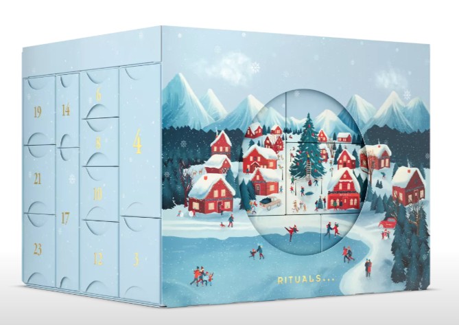 A blue Advent calendar box featuring numbered doors and an illustrated snowy village scene with red houses, people ice skating, and mountains in the background.