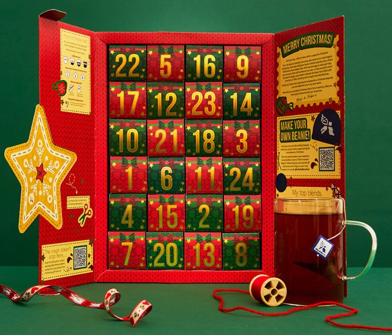 A festive Advent calendar with numbered doors is displayed alongside a glass mug of tea and holiday decorations, including ribbon, thread spool, and a star-shaped cutout.