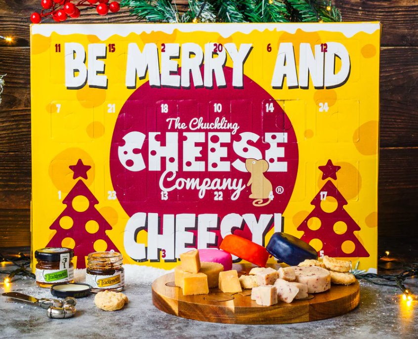chuckling cheese 2025 A festive cheese advent calendar with "Be Merry and Cheesy!" text, assorted cheeses, crackers, and spreads arranged in front on a wooden surface with holiday decorations.