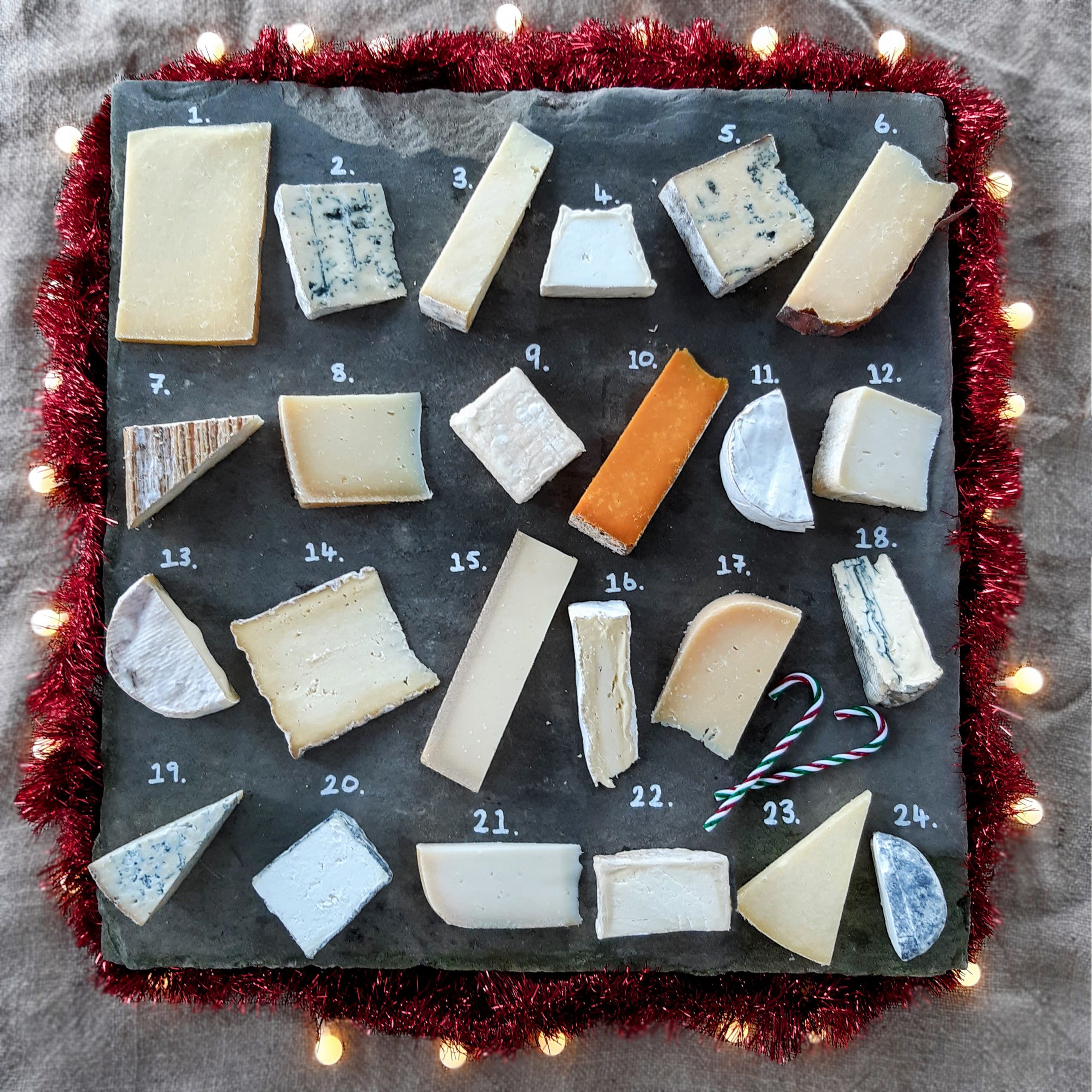 courtyard dairy 2025 A slate board with 24 assorted cheese pieces, each numbered, bordered with red tinsel and white lights, and two small candy canes placed near the lower right corner.
