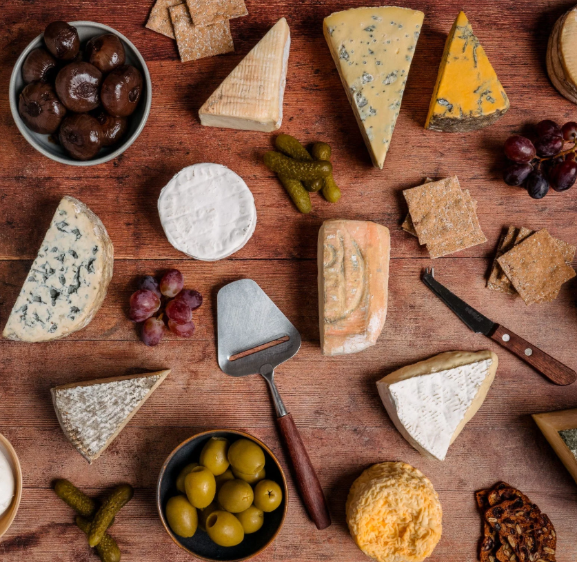 george and joseph 2025 An assortment of cheeses, crackers, pickles, olives, grapes, and mushrooms arranged on a wooden table with cheese knives.