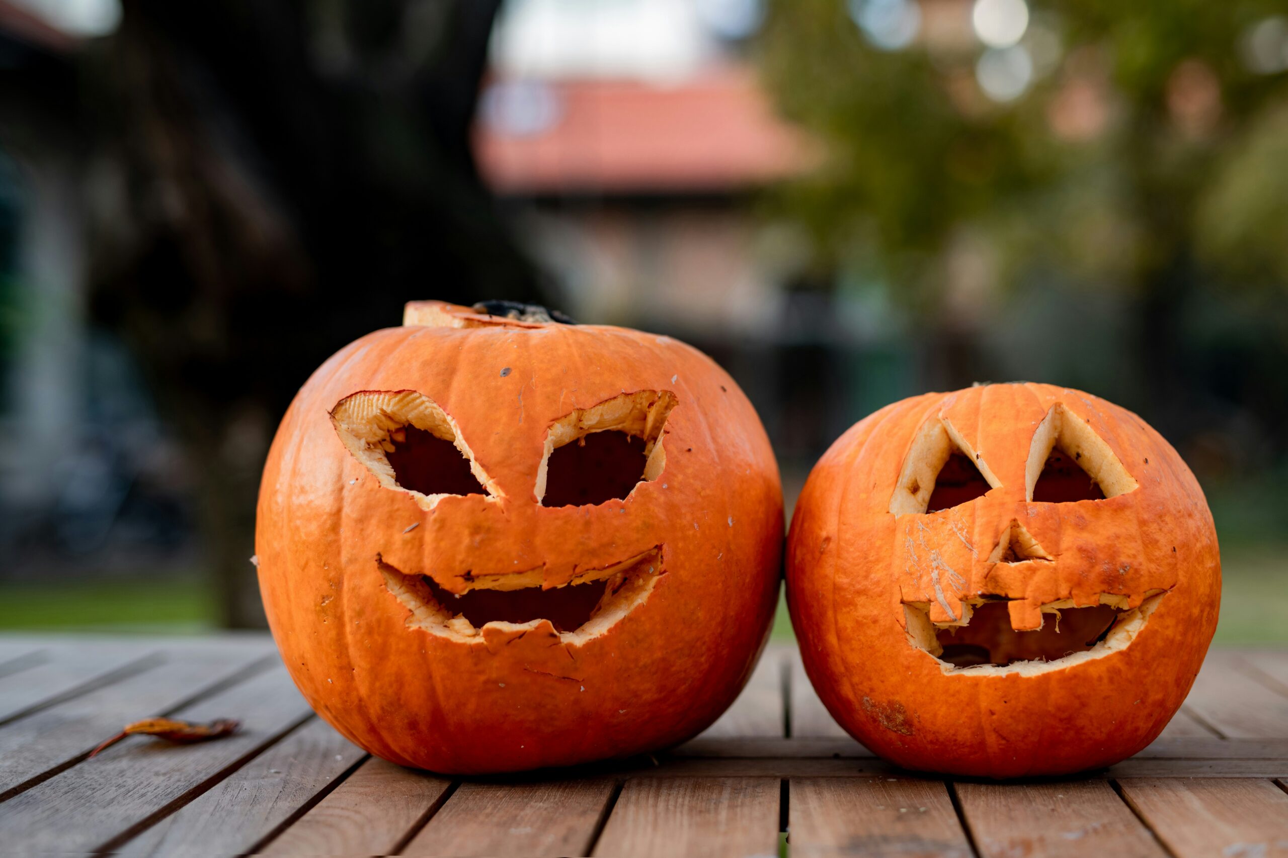 jack o lantern Two carved pumpkins with smiling faces sit side by side on a wooden table outdoors, with blurred trees and buildings in the background.