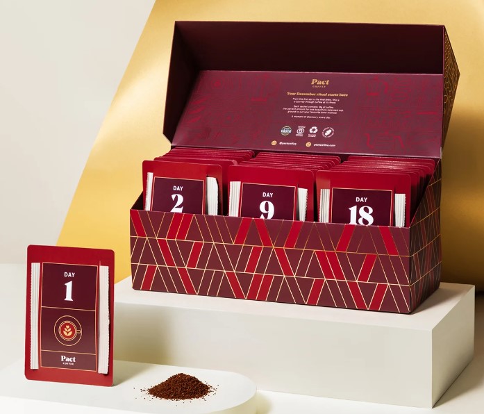 A red and gold box contains numbered coffee sachets arranged in order. In front, a card labeled "Day 1" is propped up next to a small dish of ground coffee.