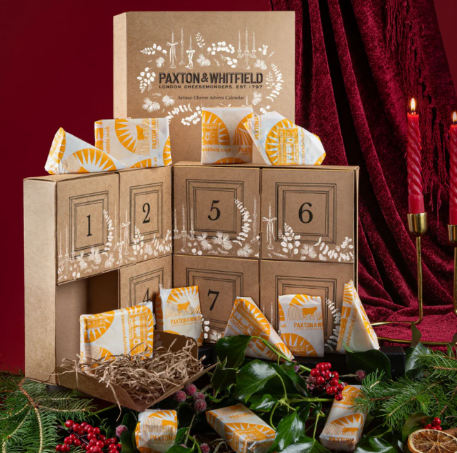 paxton and whitfield 2025 A cardboard advent calendar box labeled "Paxton & Whitfield" with numbered compartments, assorted wrapped cheeses, red candles, and festive greenery.