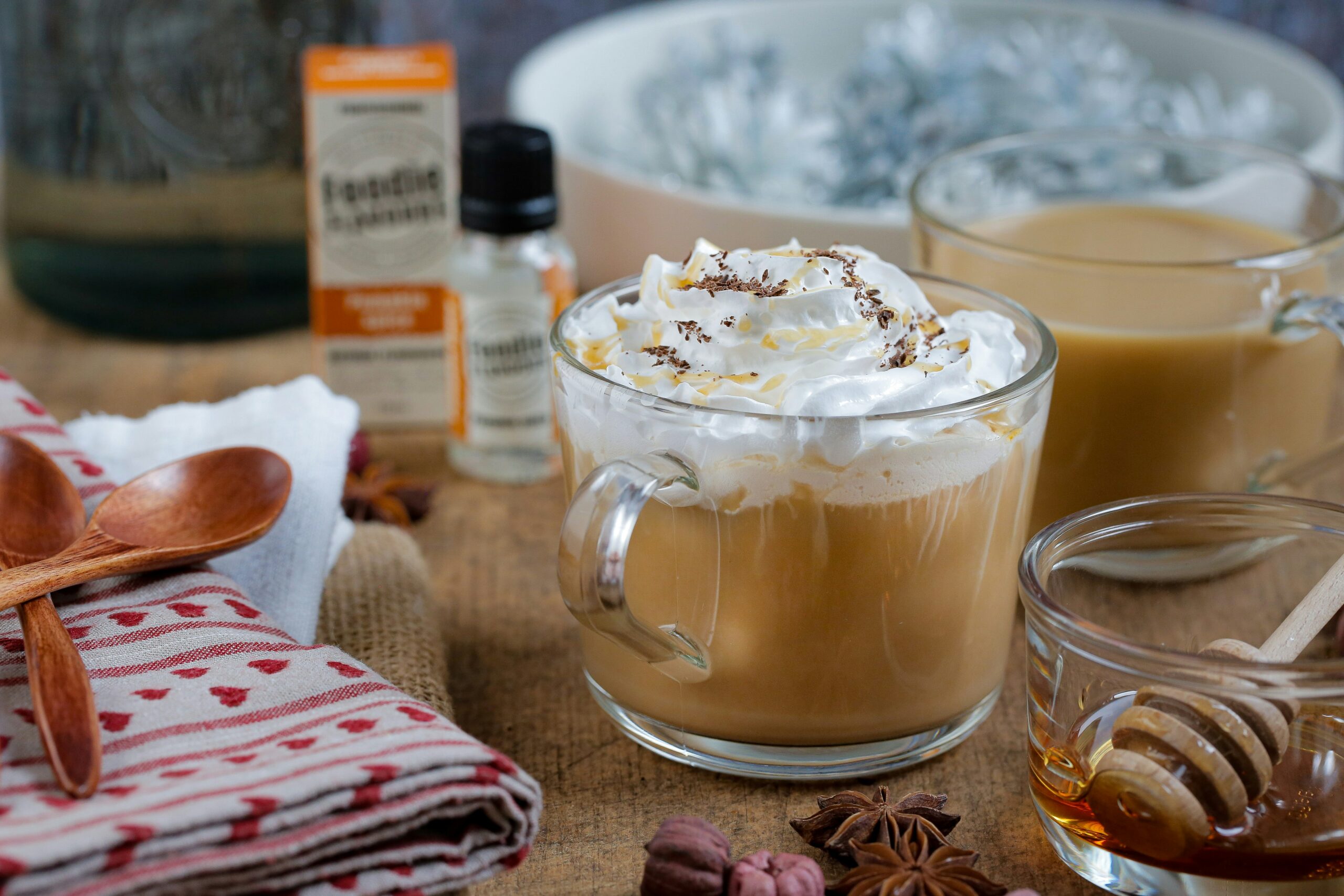 pumpkin spice latte A glass mug of coffee topped with whipped cream sits on a table with a honey dipper, a glass of coffee, wooden spoons, napkins, and a bottle of flavoring nearby.