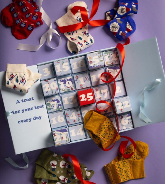 An advent calendar box with numbered compartments, surrounded by various colorful pairs of festive socks, on a purple surface.
