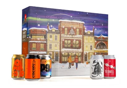 A festive box decorated with a winter town scene is shown behind five assorted cans of craft beer, each with unique labels and colors.