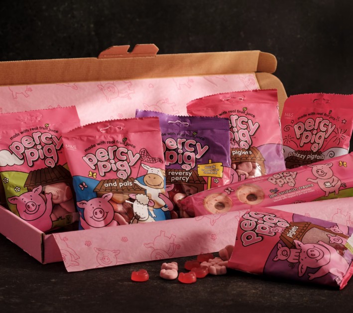 An open box displays various bags of Percy Pig gummy candies, some candy pieces scattered in front, set against a dark background.