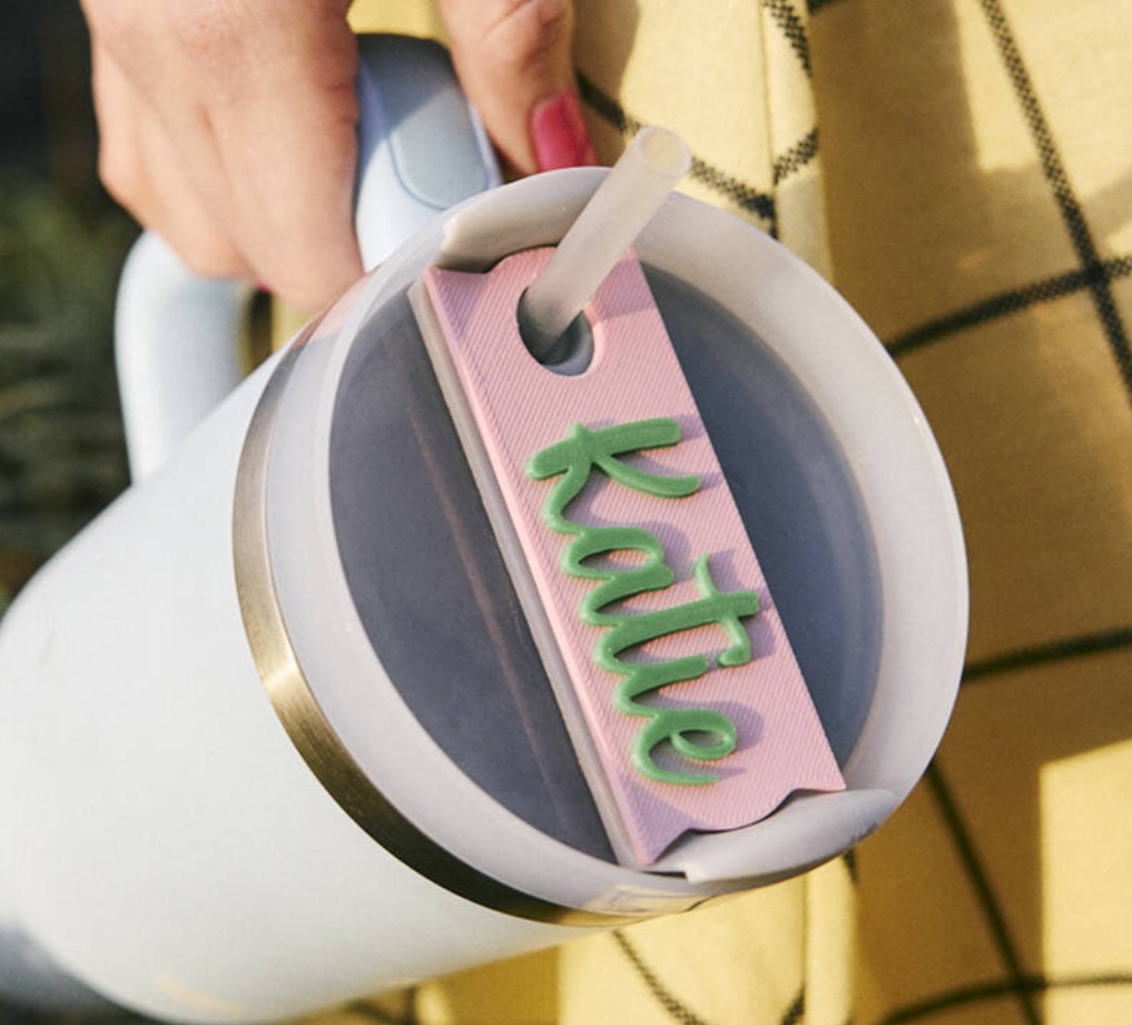 A person holds a light blue tumbler with a lid and straw. A pink name tag with "Katie" in green script is attached to the straw. The person is wearing a yellow plaid garment.