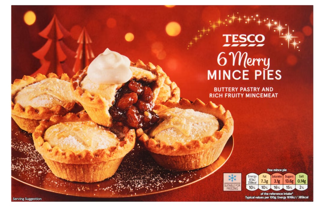 A plate of six mince pies dusted with powdered sugar, one topped with whipped cream, on a festive red background with the Tesco logo and product information.