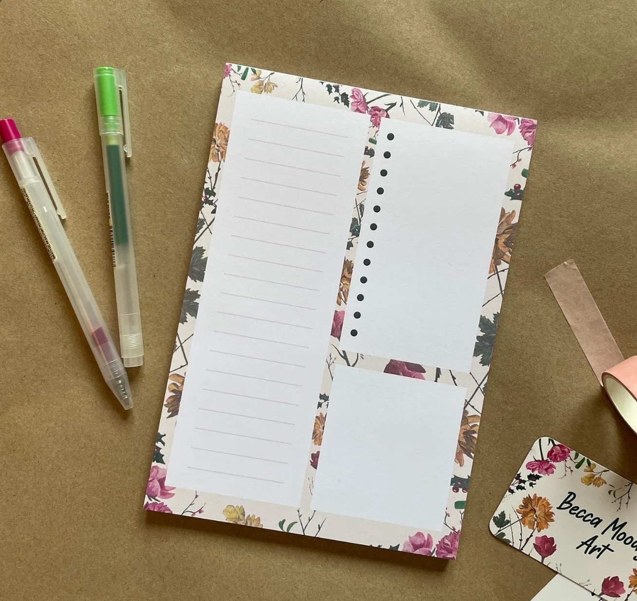 A floral notepad with three sections—one lined, one with dots, and one blank—is placed on a brown surface next to two pens and a floral business card.