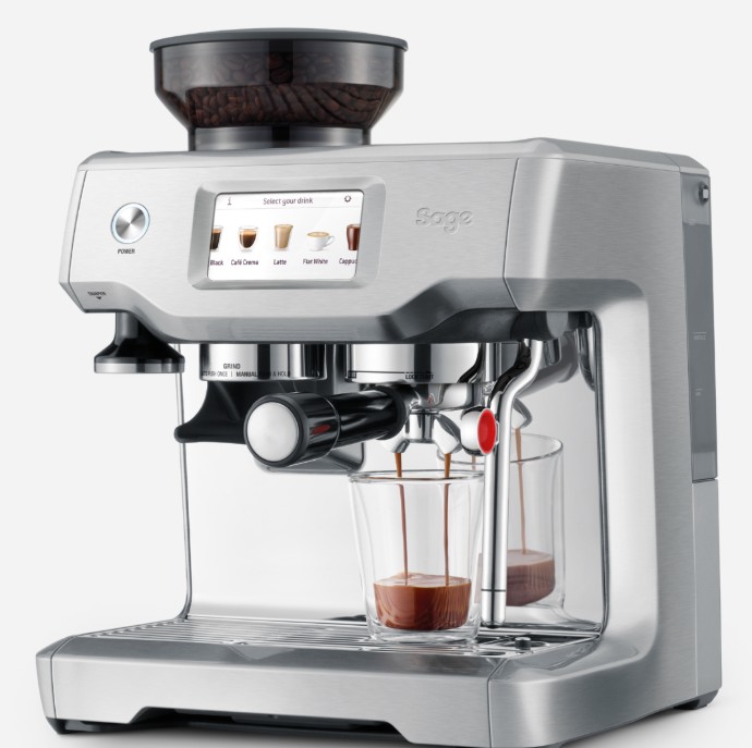 A silver Sage espresso machine with a digital display is brewing two shots of espresso into clear glass cups. Coffee beans are visible in the hopper on top.
