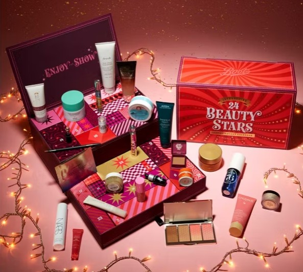 boots advent calendar A beauty advent calendar box open to reveal 24 assorted skincare and makeup products, surrounded by string lights on a pink background.