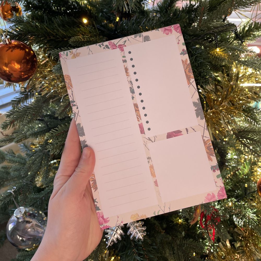 A hand holds a floral notepad with lined and blank sections in front of a decorated Christmas tree with ornaments and lights.