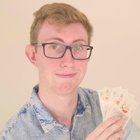 jordon cox coupon king Young man with glasses and a denim shirt holds several £10 British banknotes, fanned out in his hand, ready to save and prepare for Black Friday deals, against a plain light background.