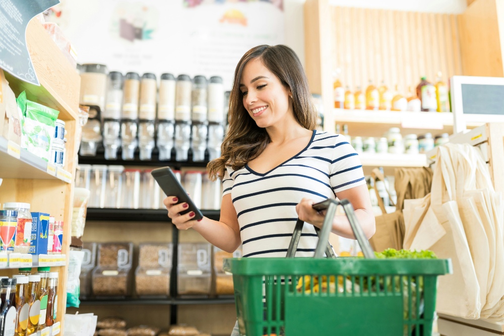 Woman holding a green shopping basket and looking at her smartphone while acting as a mystery shopper in a grocery store.
