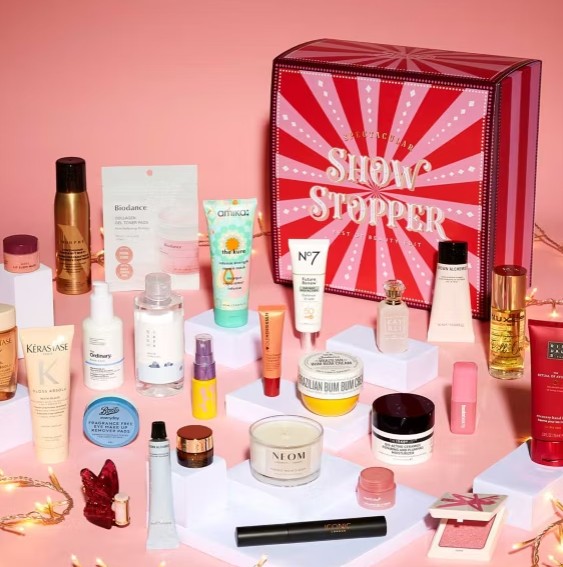 A variety of skincare, haircare, and makeup products are arranged around a red “Show Stopper” gift box on a pink background with some string lights.